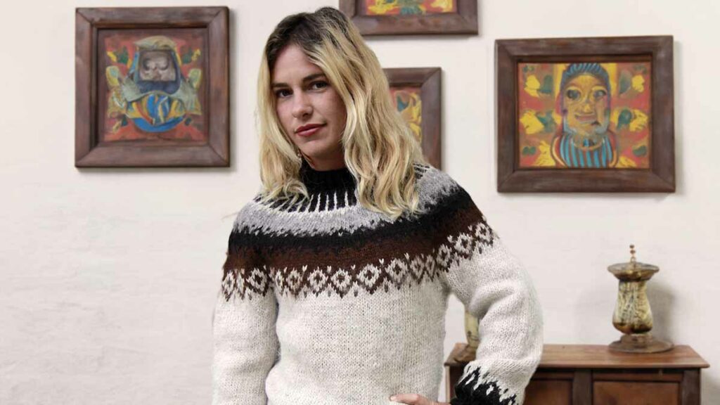 Una donna bionda indossa un maglione invernale fatto a mano con 100% lana di alpaca. Maglione di colore bianco naturale con disegno geometrico sulle spalle. 